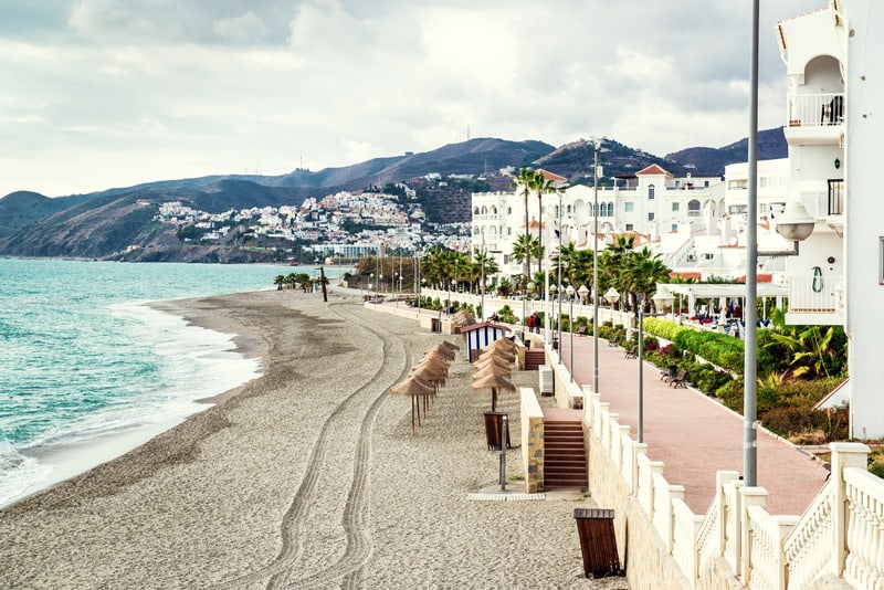 Nerja seaside with a concrete walkway on the side and a line of white houses by the beach and a view of white houses perched on a mountain