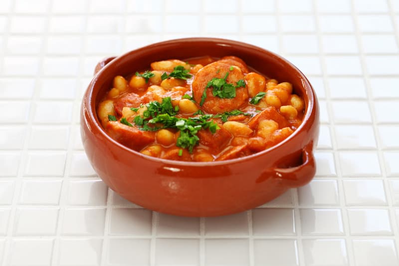 Spanish bean stew in a clay pot decorated with parsley on a white surface. 