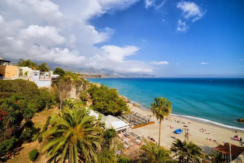 a beautiful beach filled with people with white sand and beach chairs, palm trees on the foreground, buildings on the background, and blue water and blue sky with clouds