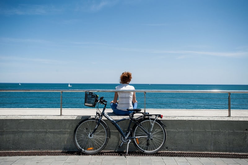 a woman sitting looking at the sea with her bike parked at her back