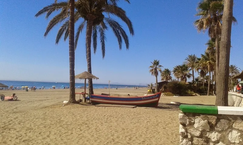 a boat in between palm trees in a beach with palm trees, people by the beach on beach umbrellas on the background