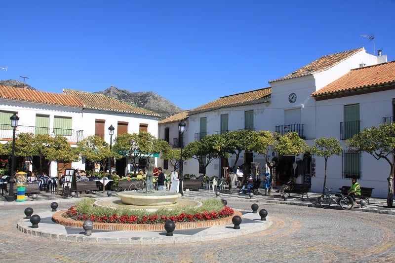 a plaza with white buildings and a fountain in the middle surrounded with trees and chairs and tables at the back