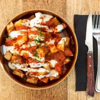 Picture of a dish of Papas bravas, made with potatoes, peppers and garlic, served on a wooden table.