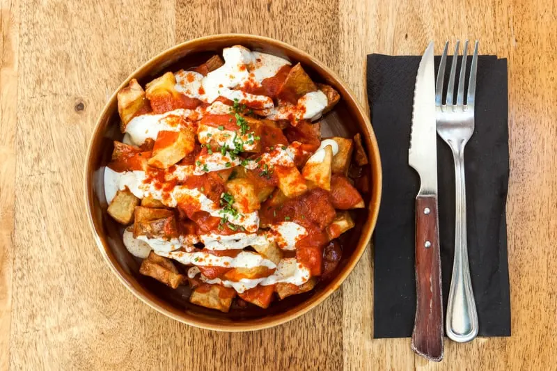 Picture of a dish of Papas bravas, made with potatoes, peppers and garlic, served on a wooden table. 