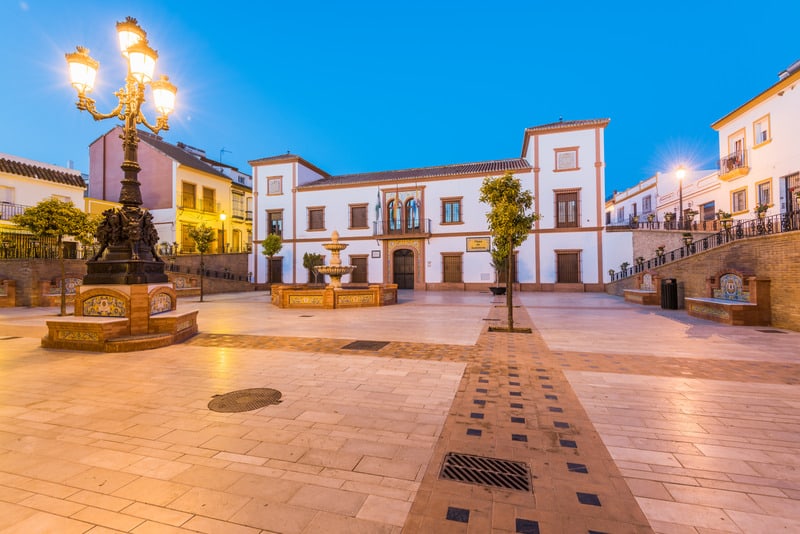 Plaza of Palos de la Frontera during the night dark sky with a white building, a water fountain, and a beautiful night city lamp