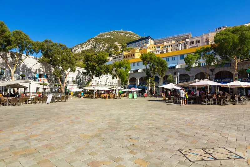 an empty plaza with tiny buildings and umbrellas on table with buildings and trees