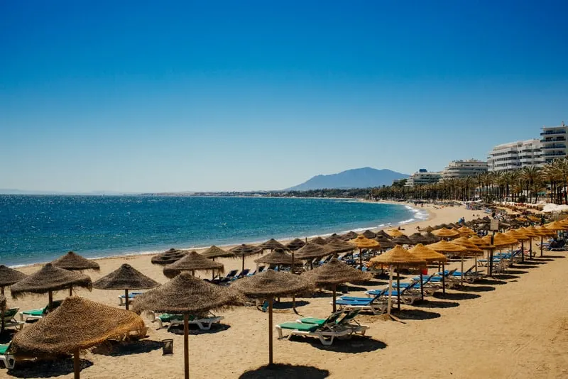 a beach in marbella filled with umbrellas and beach chairs lined  with mountains at the back