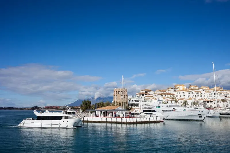 boats docked on a port with a view of white houses on a hill with clouds on the sky on a bright day