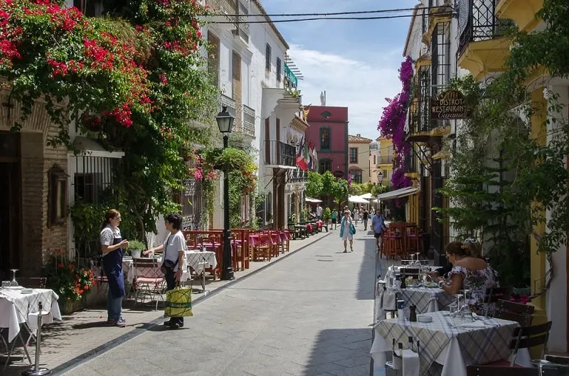 a street in old town marbella with tables and chairs on the side, people, old houses for restaurants, and flowers