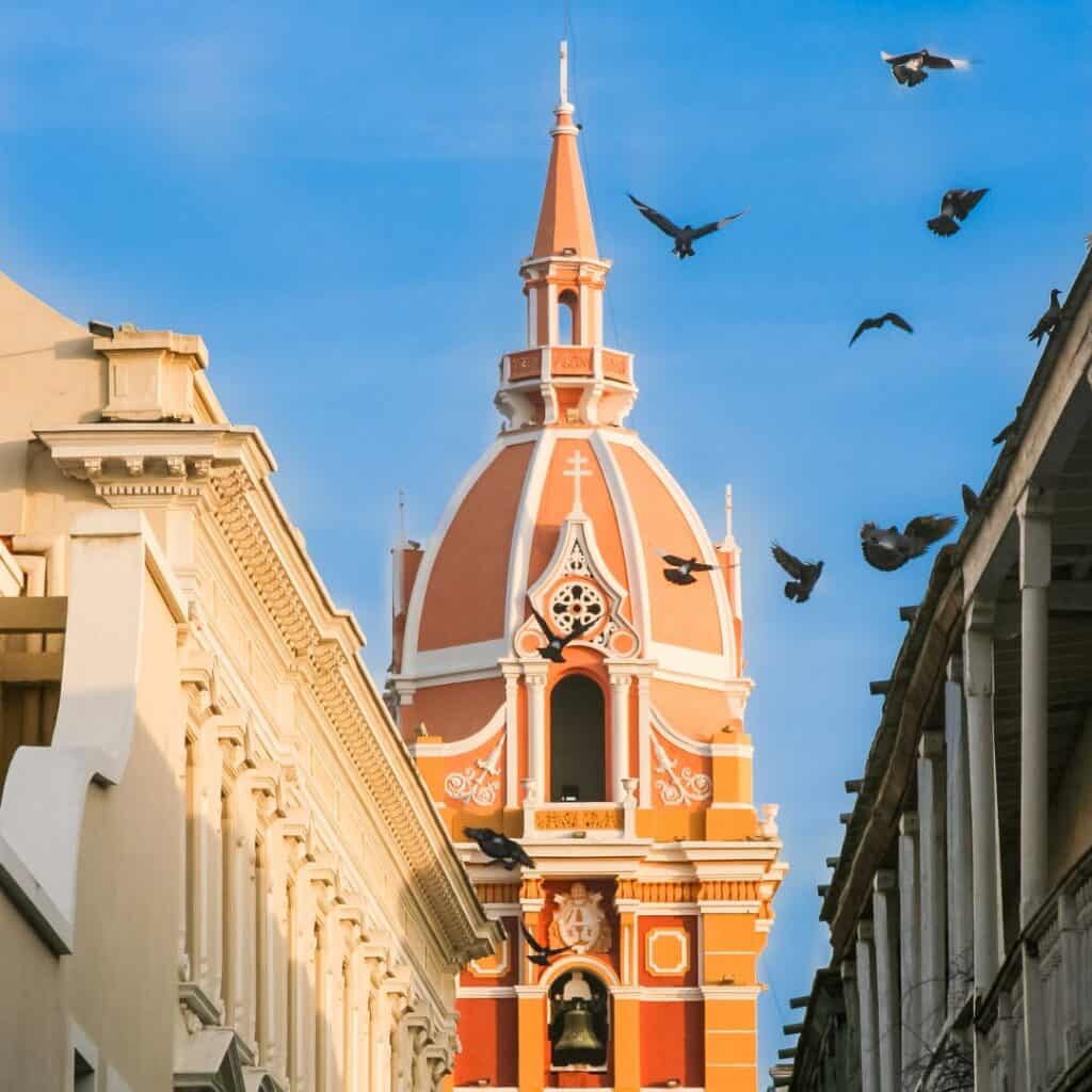 a view of a church tower between highrise buildings with birds flying in the sky