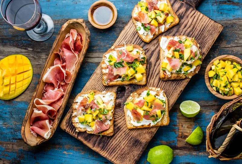 Toasts with cream cheese, ham jamon serrano and mango served on wooden board with red wine, blue wooden rustic background, top view, breakfast in spain