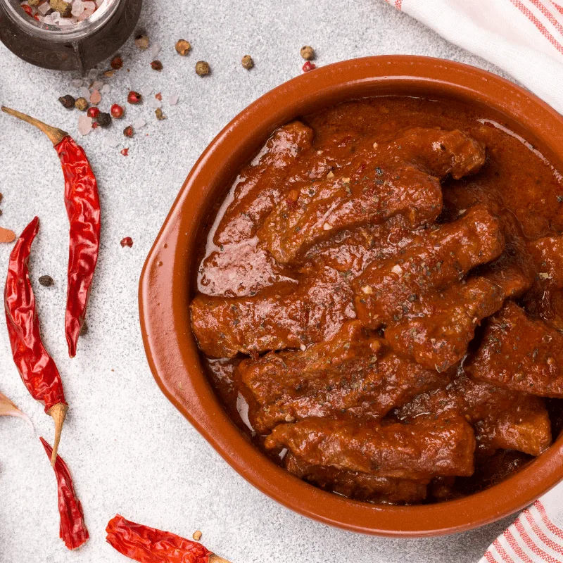 tail of a bull stewed and placed on a red bowl on a table with chili on the side of the table