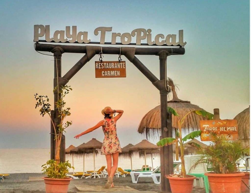 a woman posing under an archway saying Playa Tropical with beach huts and plants by the beach during a sunset