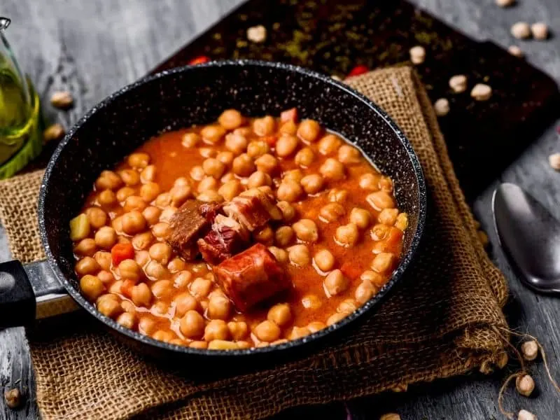 spanish garbanzo soup, red colored with beans on a black bowl on a wooden placemat and table
