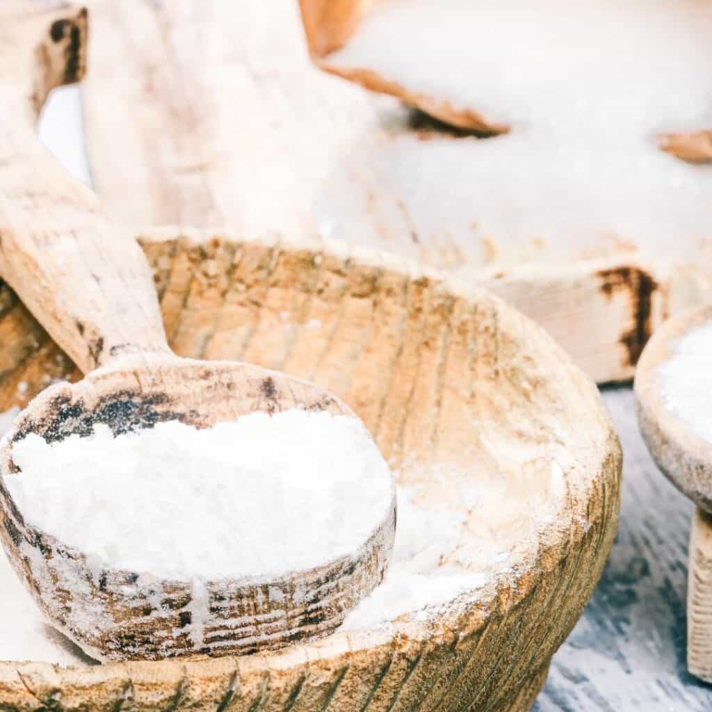 flour in a wooden spoon and bowl and sugar coming out from the bag on a wooden table for the churros cake