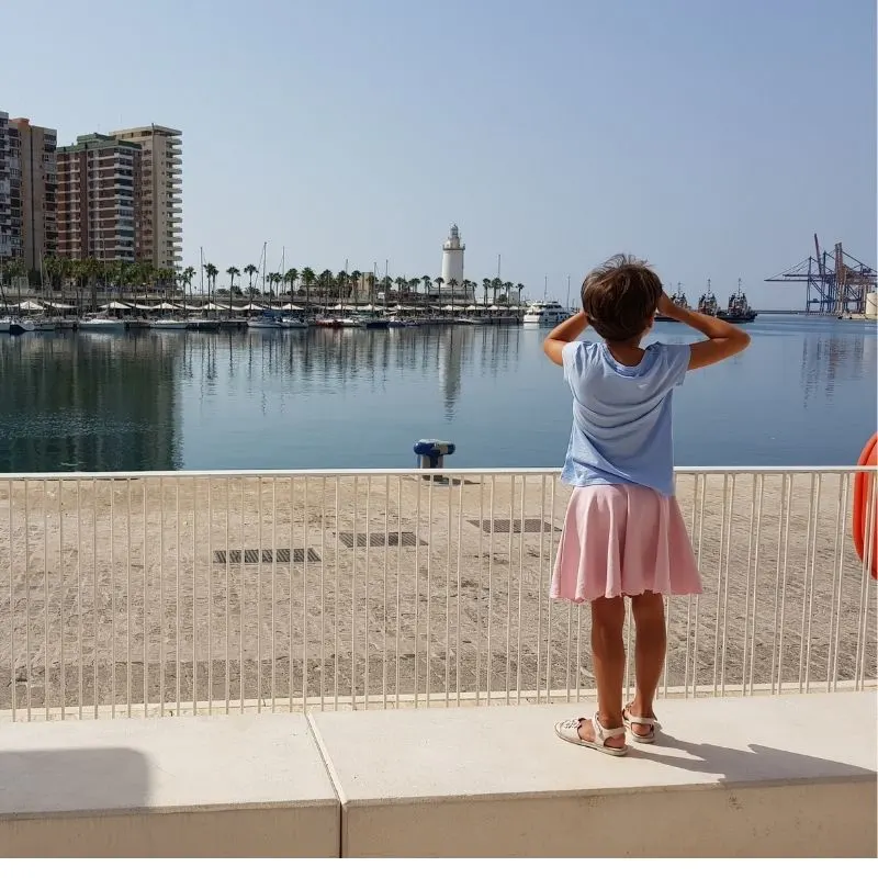 a kid looking at the  lighthouse besides palm trees and buildings across a body of water