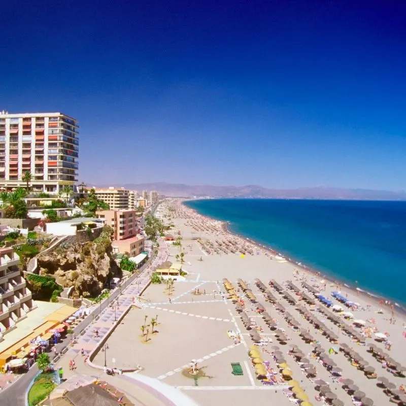 beach umbrellas and chairs lined on the beach with people walking, sunbathing, or swimming as well as buildings and resorts on the side under a blue sky