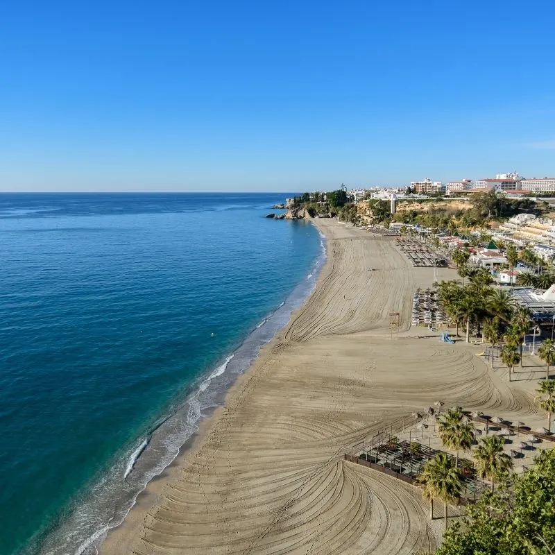 a brown sand beach with blue waters with palm trees, resorts, and beach amenities on a side