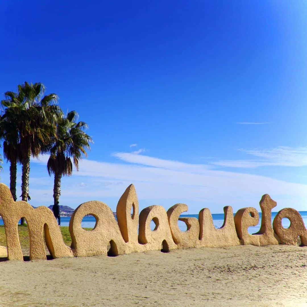 Malagueta sign made of sand with palm trees at the back on the beach on a bright blue sky, Malaga in July