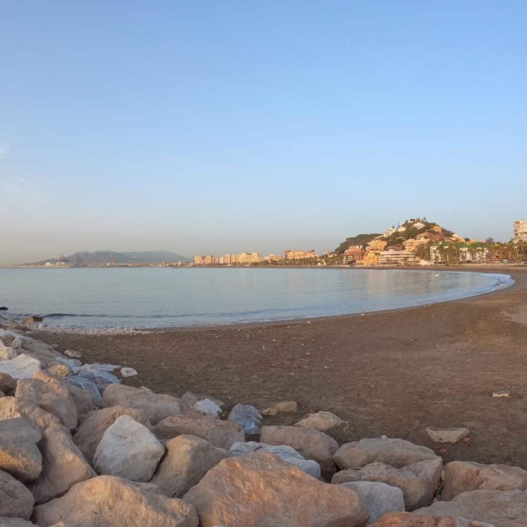 a beach with rocks and water in front of a city on a incoming sunset