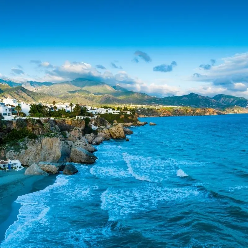 blue water splashing at the rocks and cliffs with white houses on the top with mountains on the background and a bright cloudy sky