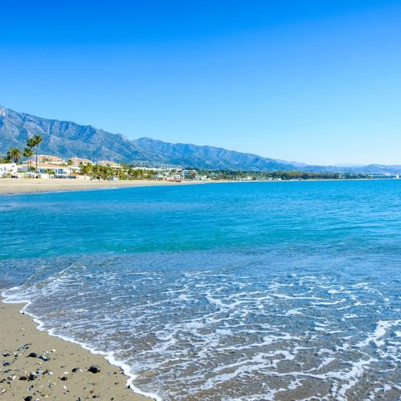 view of a blue sea and the beach in Marbella, seen from the shore with a town and mountains afar