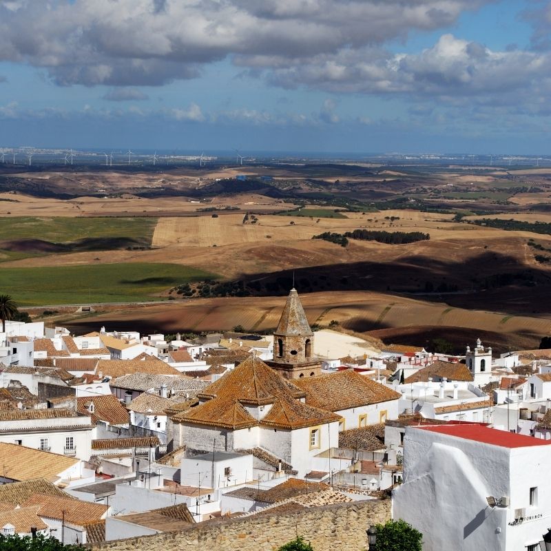 an aerial view of a town with white houses and a tower and desert and mountains at the back  with clouds on the blue sky