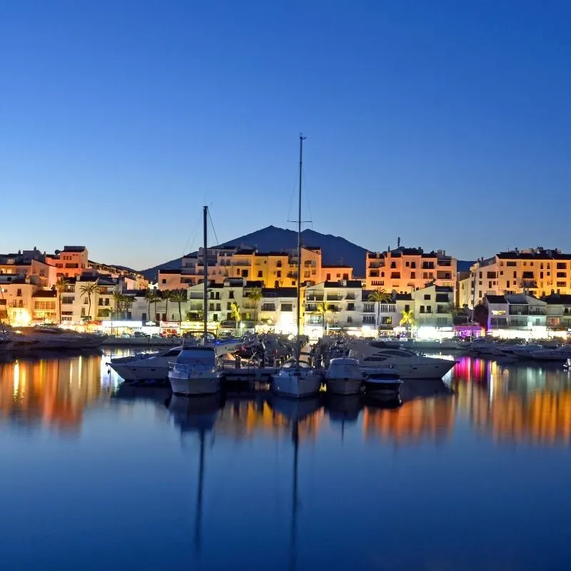 Puerto Deportivo in Marbella with boats docked as well as reflected on the river and stalls lined on the marina as well as houses and a mountain at the back at night