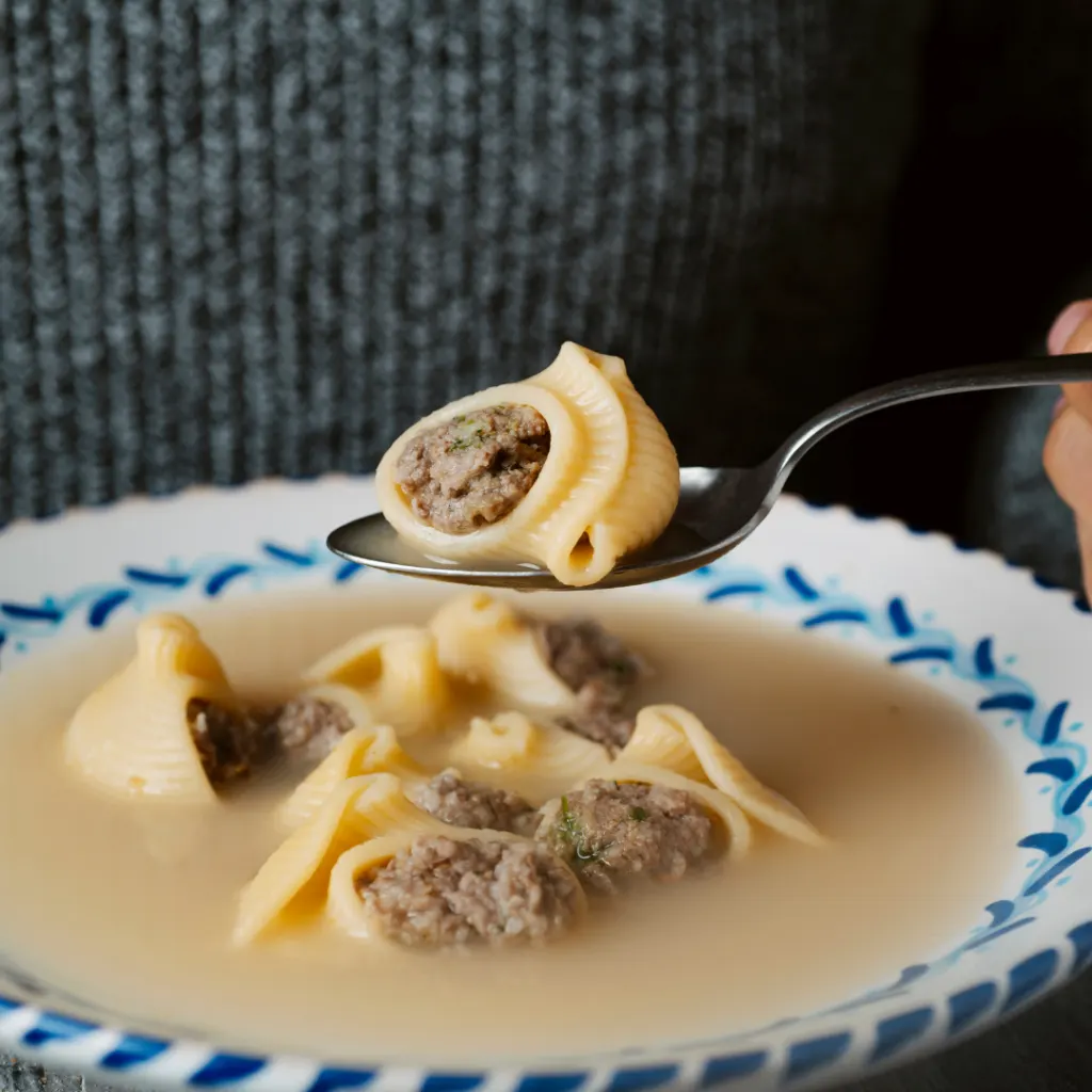 a person holding a spoon over a bowl of soup with meatballs in it