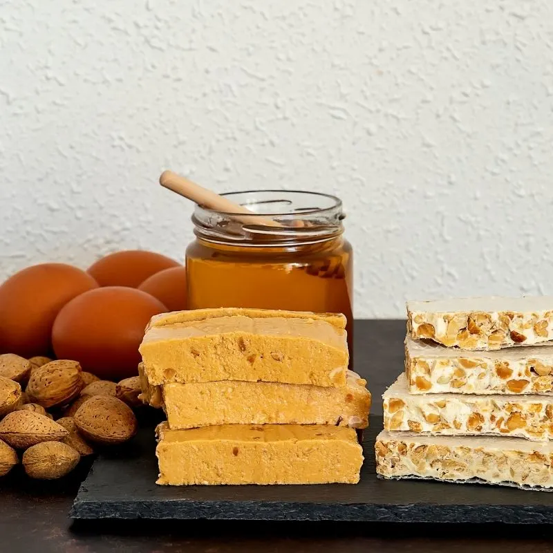 two types of turrón bars next to each other on a black plate. In the background, there's a jar of honey, 4 eggs, and some almonds. 
