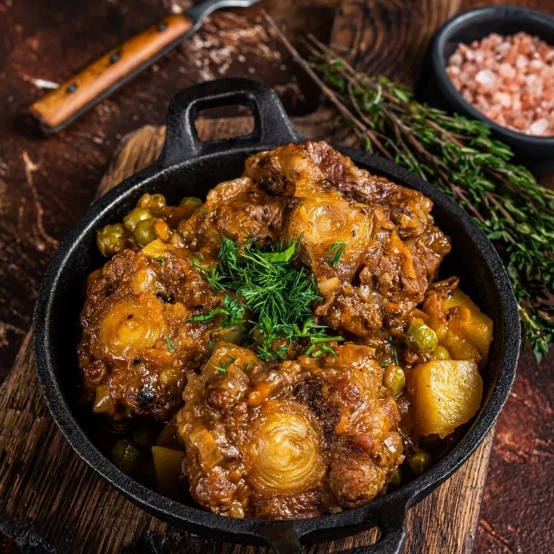 spicy oxtail recipe in a pot with herbs next to it on a wooden table