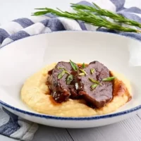 Braised pork cheeks dish in a white plate served with mashed potato on a wooden table.