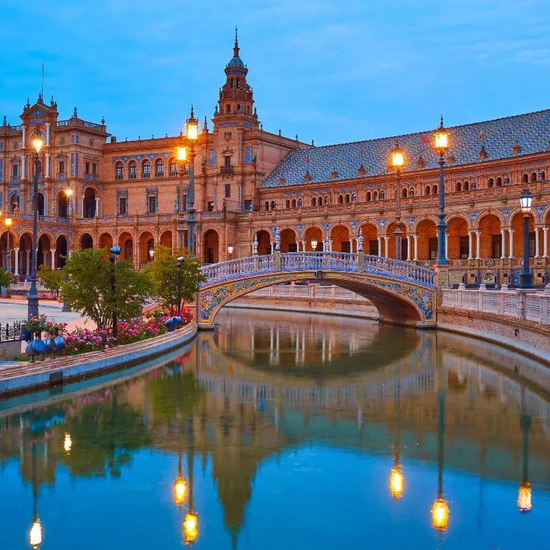 Plaza de España in Seville with a bridge over a river with lighted lamp posts and a reflection on a body of water during the night