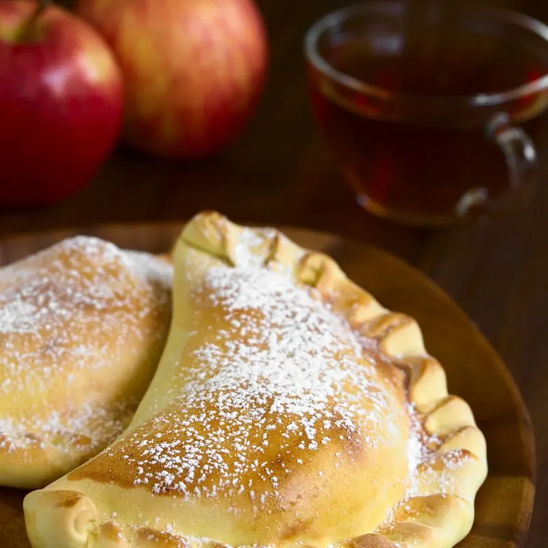 2 apple empanadas on a wooden plate, a cup of tea and 2 apples in the background. 