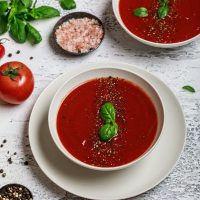 gazpacho in 2 bowls with fresh tomato and basil next to them