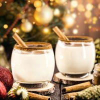 coquito drink in two glasses, decorated with two sticks of cinnamon, on a wooden table with a festive background.