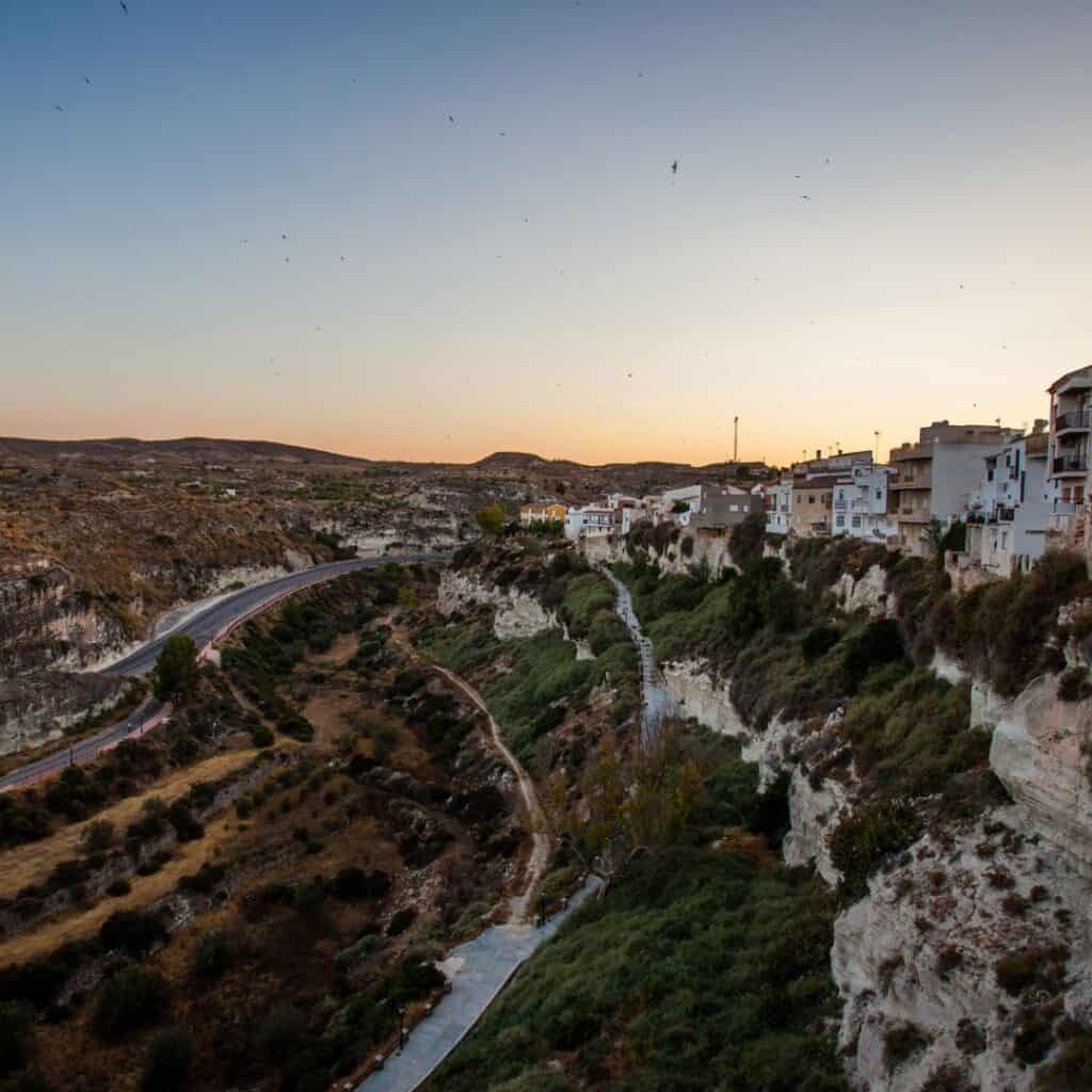 the view from the top of a hill at sunset with a town by the cliff