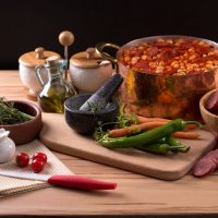 Spanish baked beans in a pot surrounded by ingredients such as tomatoes, chorizo, peppers and spices on a wooden table.