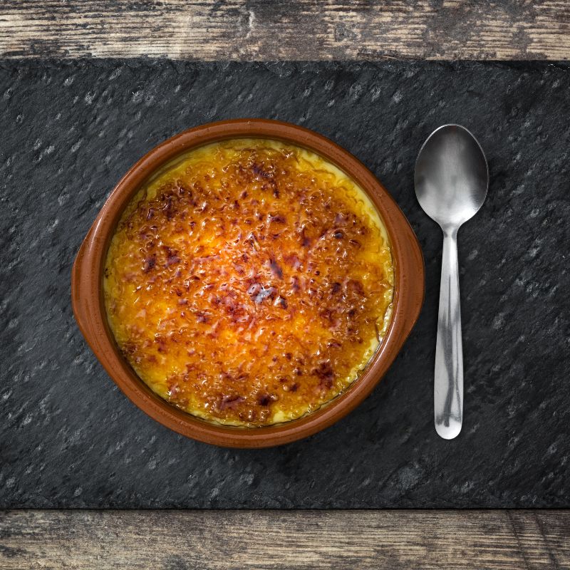 crema catalana dessert in a clay pot, on a wooden table and next to it there's a spoon. Delightful Crema Catalana Recipe