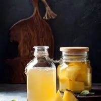 Tepache de pina in 2 jars and pineapple slices on a kitchen table.
