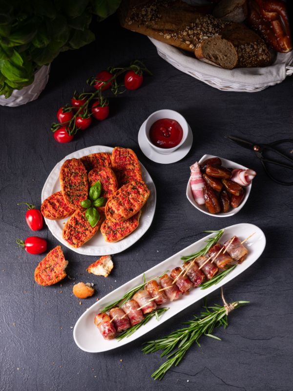 dates wrapped in bacon on a serving plate, next to tomato toast, tomatoes, dates and bread on a table.