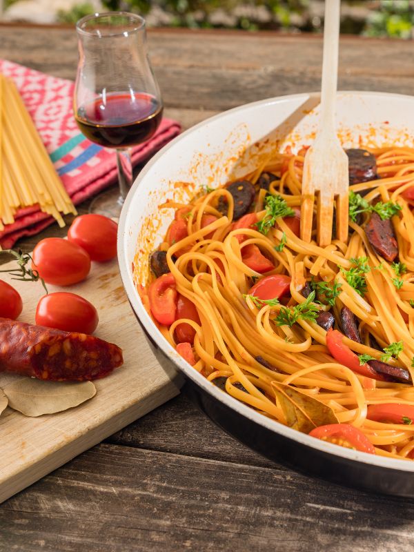 pasta with chorizo in a pan next to tomatoes and a glass of wine.