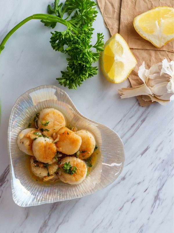 spanish scallops in a bowl with lemon garlic and parsley next to it. - Delicious Spanish Scallops with Garlic Recipe
