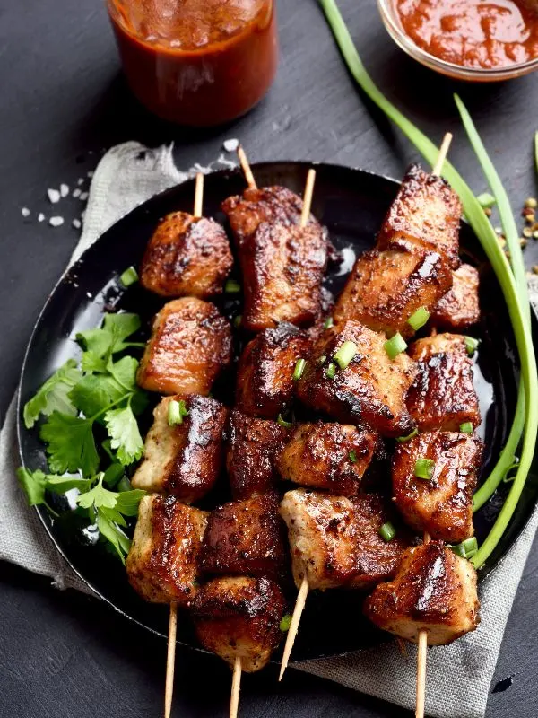 Spanish pork skewers on a black plate with red sauce next to them.