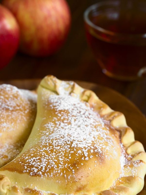 2 apple empanadas on a wooden plate, a cup of tea and 2 apples in the background.