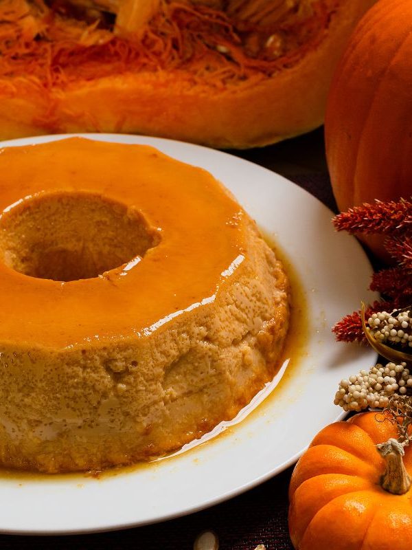 pumpkin spice flan on a white plate next to various-sized pumpkins and a dry flower bouquet.