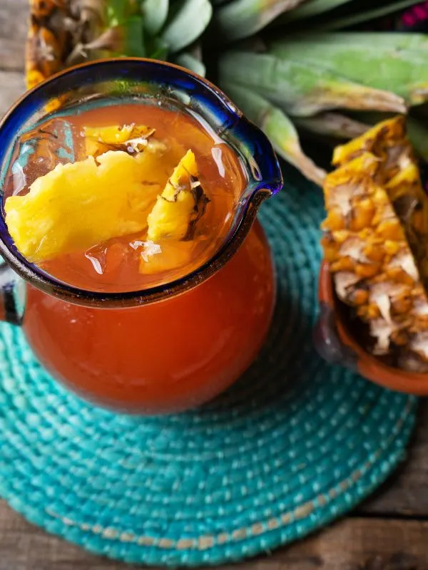 Tepache de Piña drink in a glass container with pineapple chunks next to it.