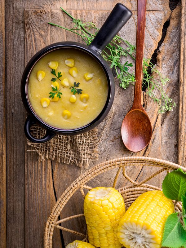 corn gazpacho in a black bowl, on a wooden table with a basket of corn next to it.