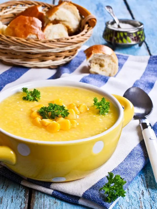 sweet corn gazpacho served in a yellow bowl with bread in the background on a blue surface.