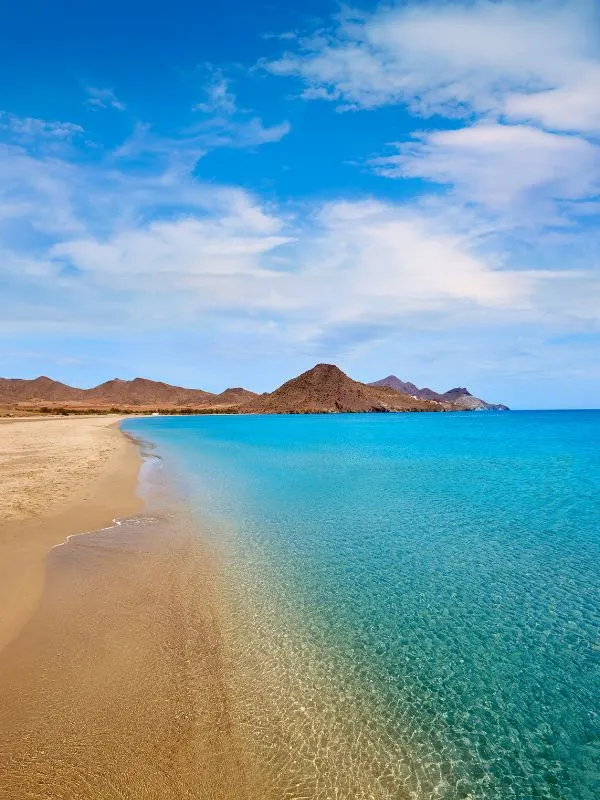 golden sand beach with turquoise waters and hills and mountains on the back under a blue sky with white wispy clouds